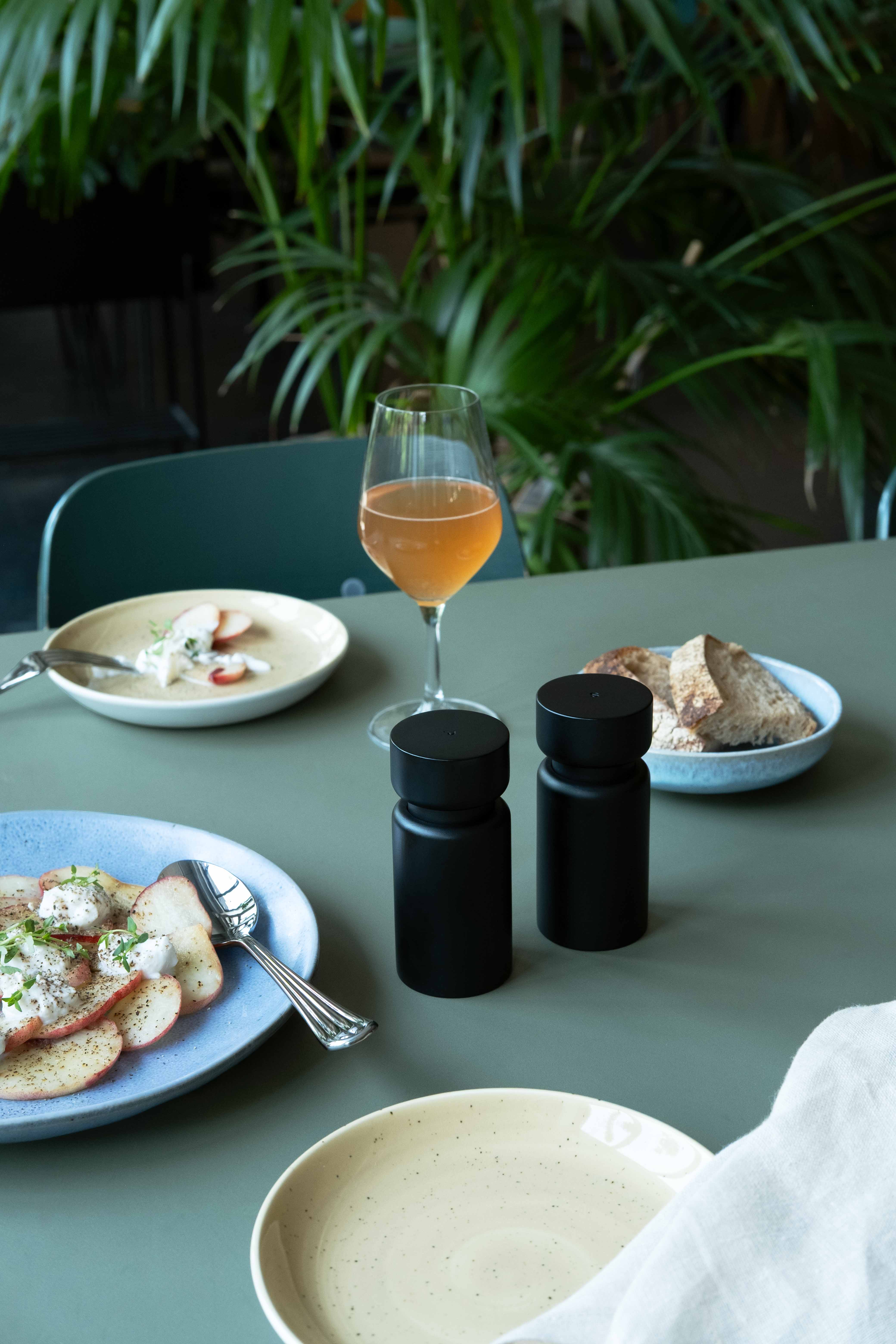 Dining table with black salt and pepper mills, sliced apples with cream, bread, and a glass of amber drink.