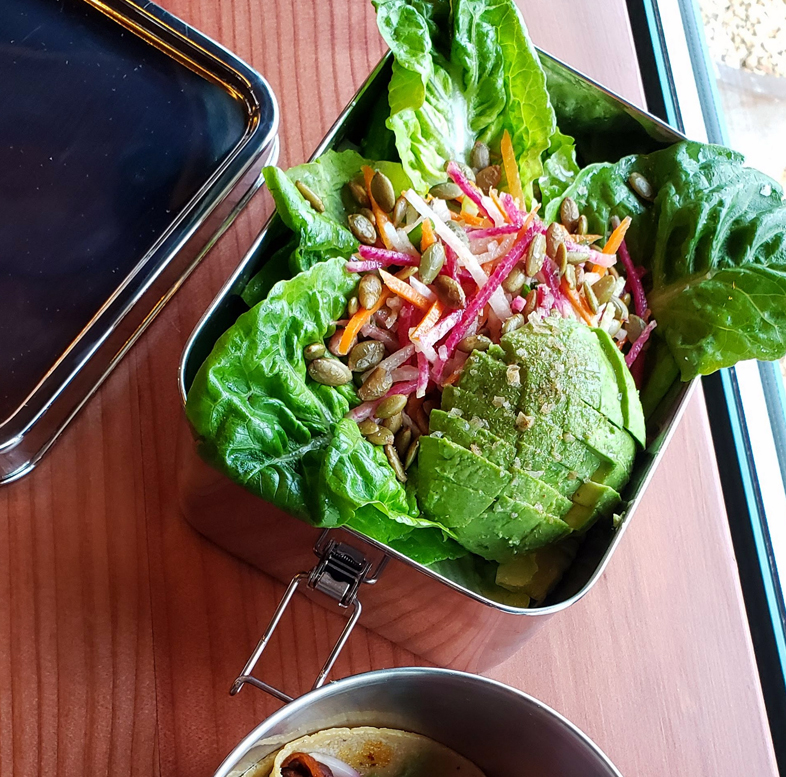Metal lunchbox with leafy greens, shredded carrots, radishes, pumpkin seeds, and sliced avocado.