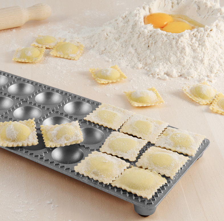 Freshly made ravioli dusted with flour on a metal tray beside flour and egg yolks.
